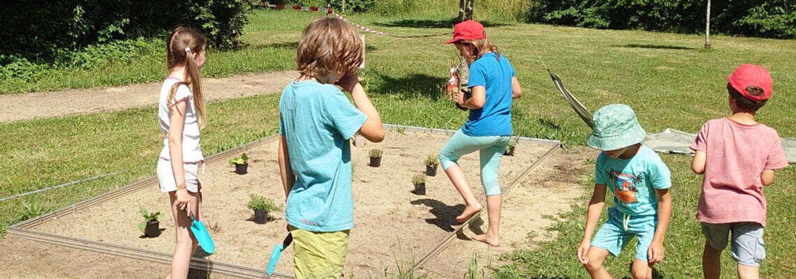 Kinder stehen um eine Sandfläche in einer parkähnlichen Umgebung. Neben ihnen stehen Blumentöpfe zum Einpflanzen.