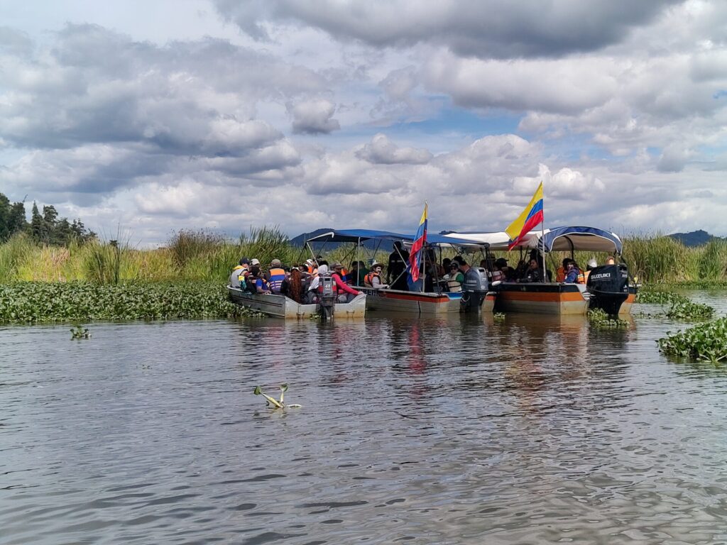 Blick auf die Flachwasserlagune Fuquene in Kolumbien. Am Ufer sind Wasserhyazinthen zu sehen sowie drei Botte mit Menschen. Die Boote sind mit der kolumbinischen Flagge geschmückt.