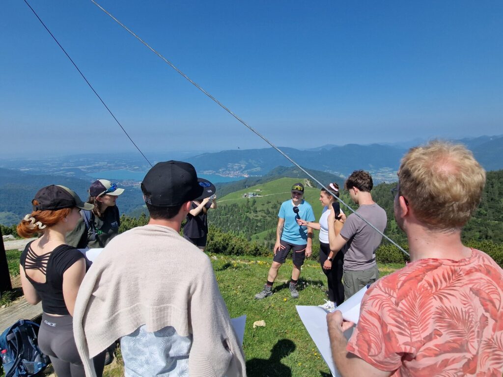 Blick von einem Berggipfel auf eine offene Landschaft mit See und Bergen. Im Vordergrund stehen junge Menschen in Wanderkleidung. Eine junge Frau hält einem Wanderer ein Mikrofon hin, ein junger Mann filmt die Szene mit dem Smartphone.