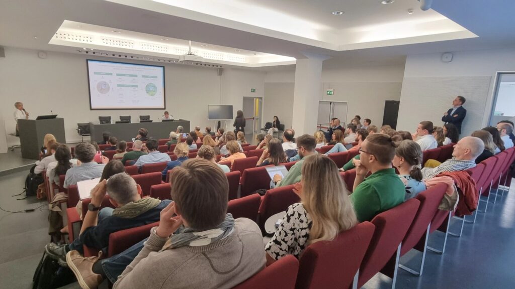 Aus leichter Vogelperspektive sieht man den Konferenzraum. Die Zuhörer*innen sitzen auf dunkelroten Stühlen. Auf der Leinwand ist schon eine Präsentation zusehen, vor der vier Stühle stehen, wobei nur der rechte von einem Mann besetzt ist. Die Decke wirddurch indirekte Beleuchtung mit weißem Licht angestrahlt. Der Raum wirkt hell und freundlich.