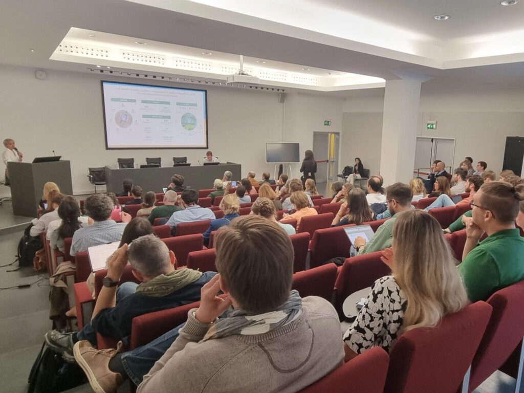 Aus leichter Vogelperspektive sieht man den Konferenzraum. Die Zuhörer*innen sitzen auf dunkelroten Stühlen. Auf der Leinwand ist schon eine Präsentation zusehen, vor der vier Stühle stehen, wobei nur der rechte von einem Mann besetzt ist. Die Decke wirddurch indirekte Beleuchtung mit weißem Licht angestrahlt. Der Raum wirkt hell und freundlich.