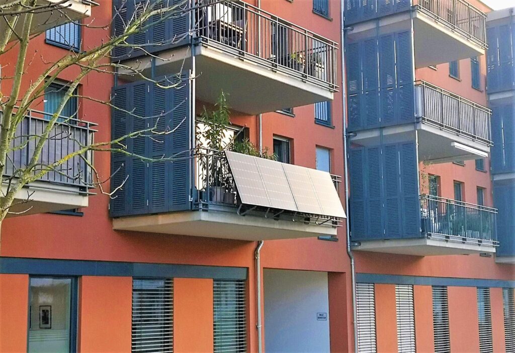 Zusehen ist ein Wohnkomplex mit Balkonen. Die Farbe des Hauses ist Ocker Rot. Der Blick fällt auf einen Balkon, bei dem PV-Anlagen angebaut wurden.