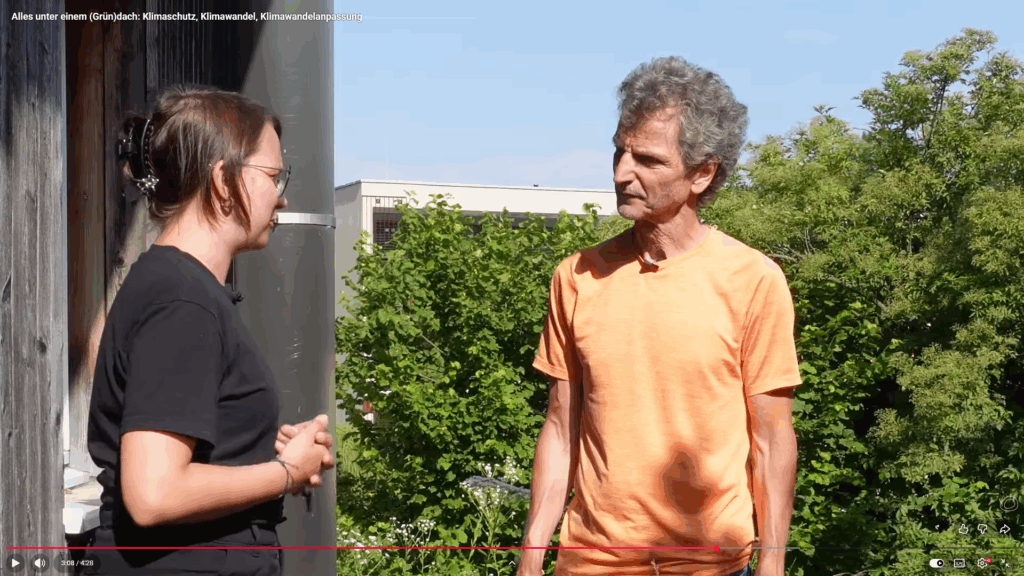 Ewelina Langer vom Energieinstitut Vorarlberg interviewt Martin Staudinger, Experte für Haustechnik, warum er vor 28 Jahren ein Haus mit Dachbegrünung erworben hat. Das Bild ist ein Screenshot aus dem YouTube Video des Interviews. Beide stehen sich gegenüber. Frau Langer steht links in einem schwarzen T-Shirt und Herr Staudinger steht rechts in einem orangenen T-Shirt. Im Hintergrund sieht man Grüne Bäume.