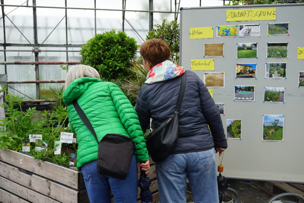 Zu sehen sind zwei Frauen, die mit dem Rücken zum Bild stehen. Vor Ihnen ist eine Pinnwand mit der Aufschrift: Zukunftsgrün, an der Naturereignisse wie Hitze und Trockenheit fotografisch festgehalten sind. Rechts Daneben steht ein Kräuterbeet.