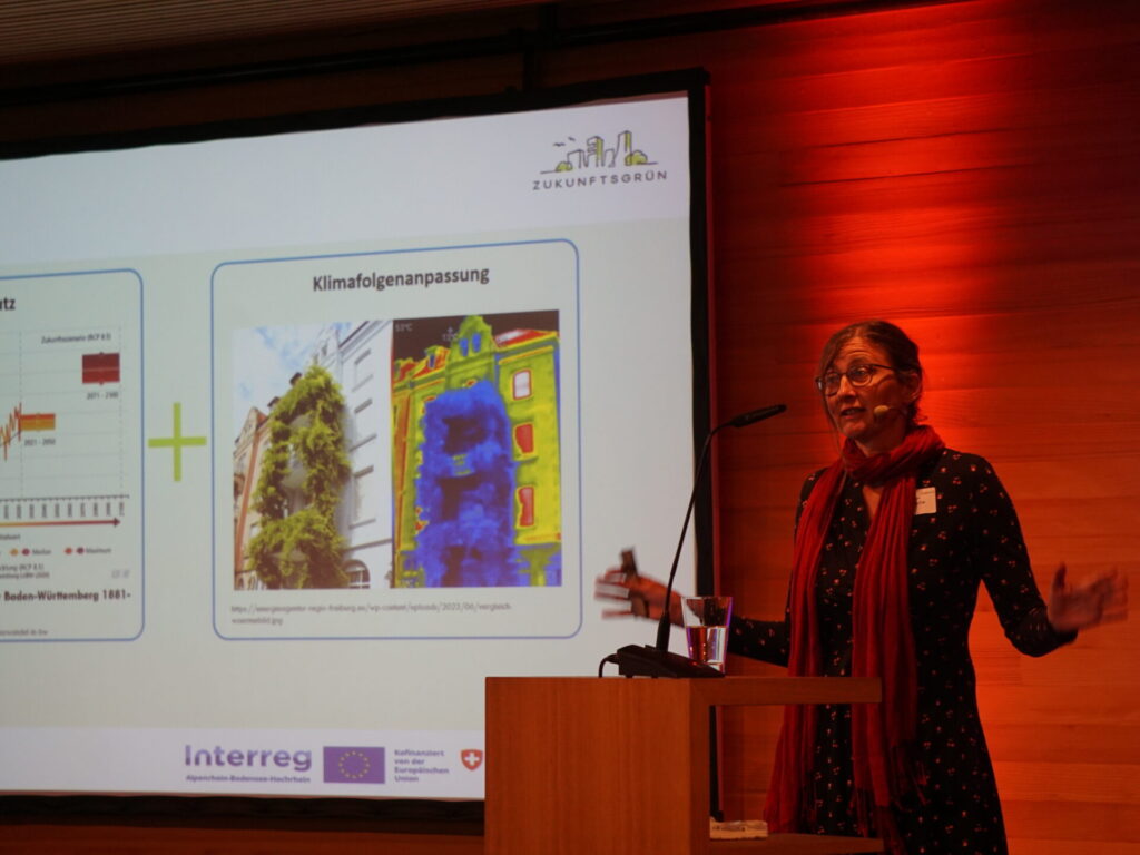 Daniela Dietsche steht auf dem Podium und präsentiert ihren Vortrag. Sie stellt die Kombination von Gründach und PV als Multitalent für die Energiewende vor. Hinter ihr wirft ein Scheinwerfer ein Lichtstrahl in rot an die Wand.