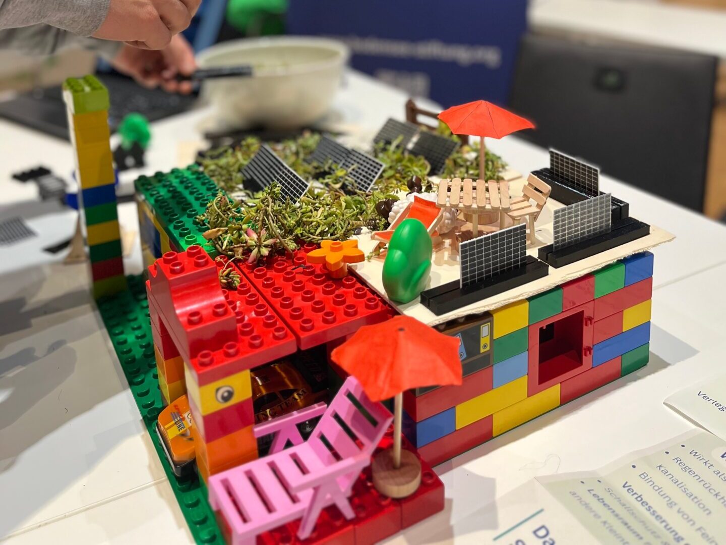 Das Lego Haus mit Gründach und PV-Anlage. Davor die Garage mit Auto, dem Sonnenschirm und Dachterrasse.