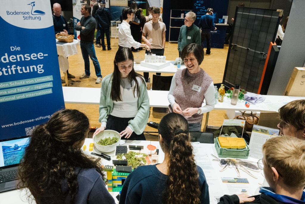 Links steht das Banner der Bodensee-Stiftung. Auf dem Tisch daneben ist ein Lego Haus mit Gründach und PV-Anlage aufgebaut worden. Die Schulkinder stehen davor und basteln daran rum.