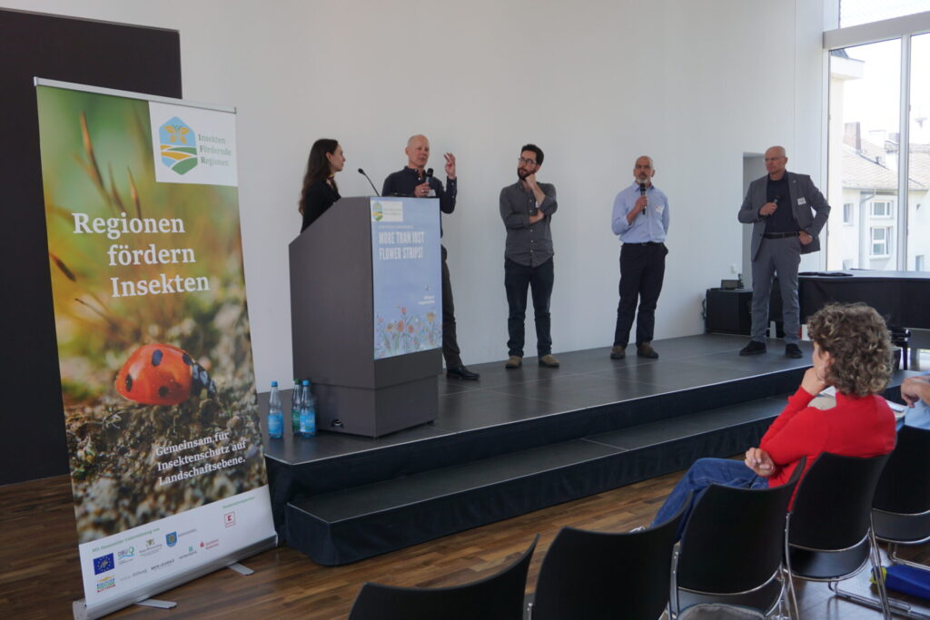 Im Blick sind 5 Personen auf dem Podium, die über verschiedene Themen diskutieren. Patrick Trötschler hält das Mikrofon. Links sieht man den Roll-Up vom Projekt Insektenfördernde Regionen.