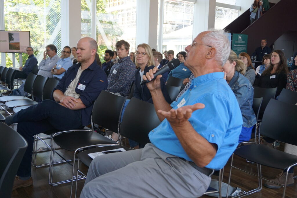 Zusehen ist ein älterer Herr in einem hellblauen Hemd, der ein Mikrofon hält. Hinter ihm sitzen viele Zuhörer*innen.