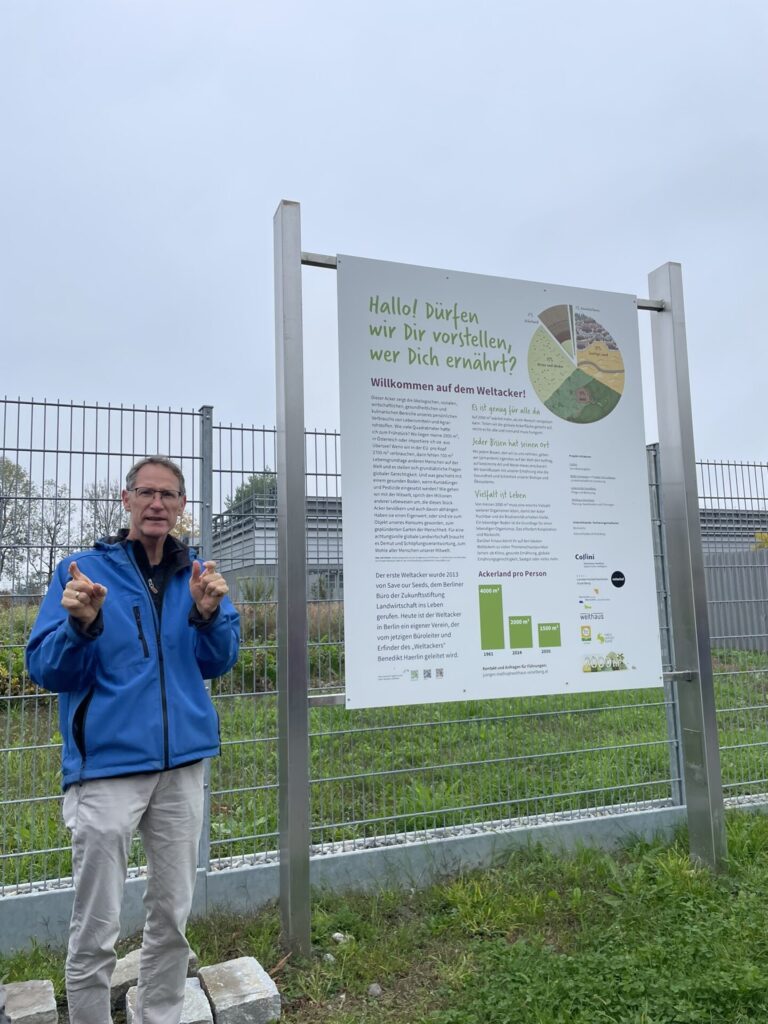 Jürgen Mathis steht links vor dem Infoplakat und erklärt den Teilnehmer*innen was der Weltacker ist.