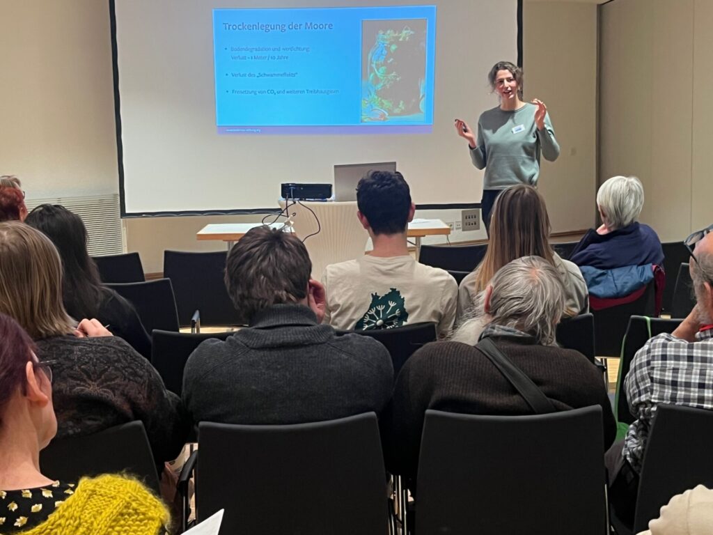 Luisa Gedon referiert vor einer Leinwand und einem zuhörenden Publikum, das in Kinobestuhlung vor ihr sitzt.