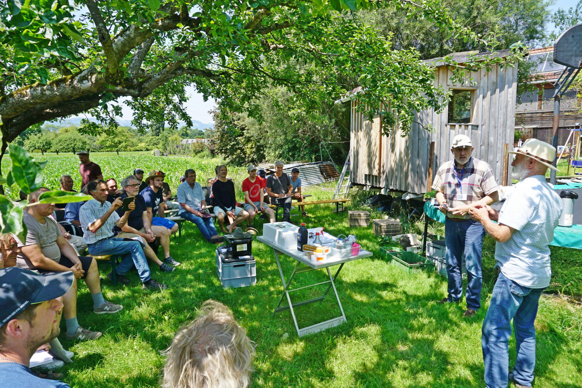 Eine Gruppe von Menschen sitzt im Freien unter einem Baum im Halbkreis auf Bänken. Die Menschen blicken auf zwei Männer, die in der Mitte an einem Tisch stehen und referieren.