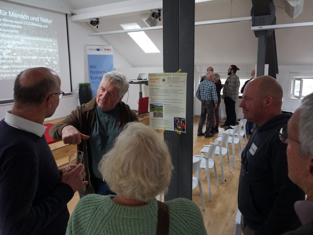 In einem Tagungsraum stehen miteinander diskutierend zwei Gruppen jeweils um Pfosten, an denen Poster hängen.