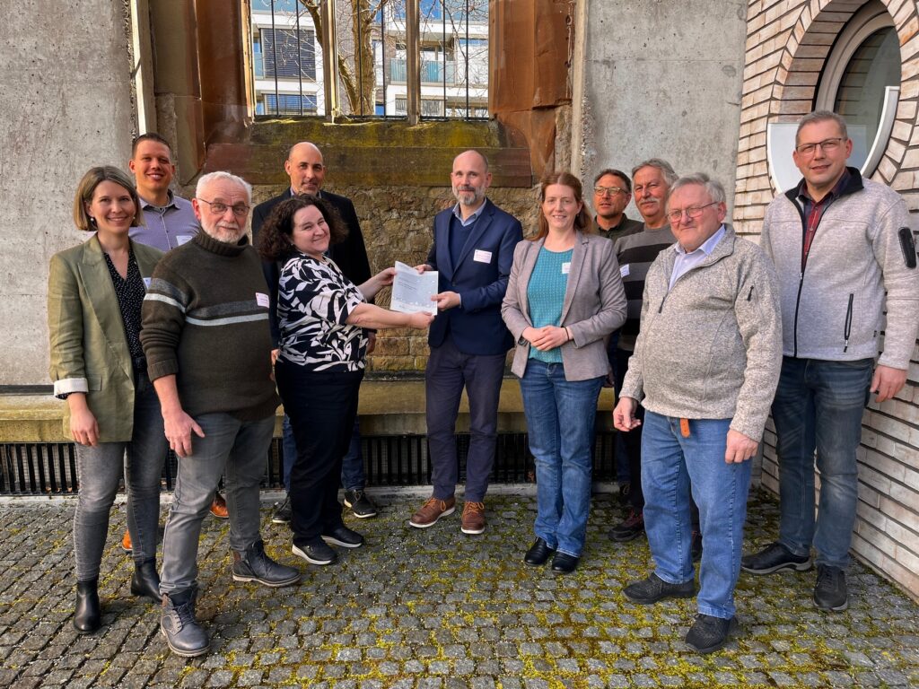 Eine Gruppe von elf Menschen, Männern und Frauen, blickt in die Kamera. In der Bildmitte übergibt eine Frau eine Publikation an einen Mann.