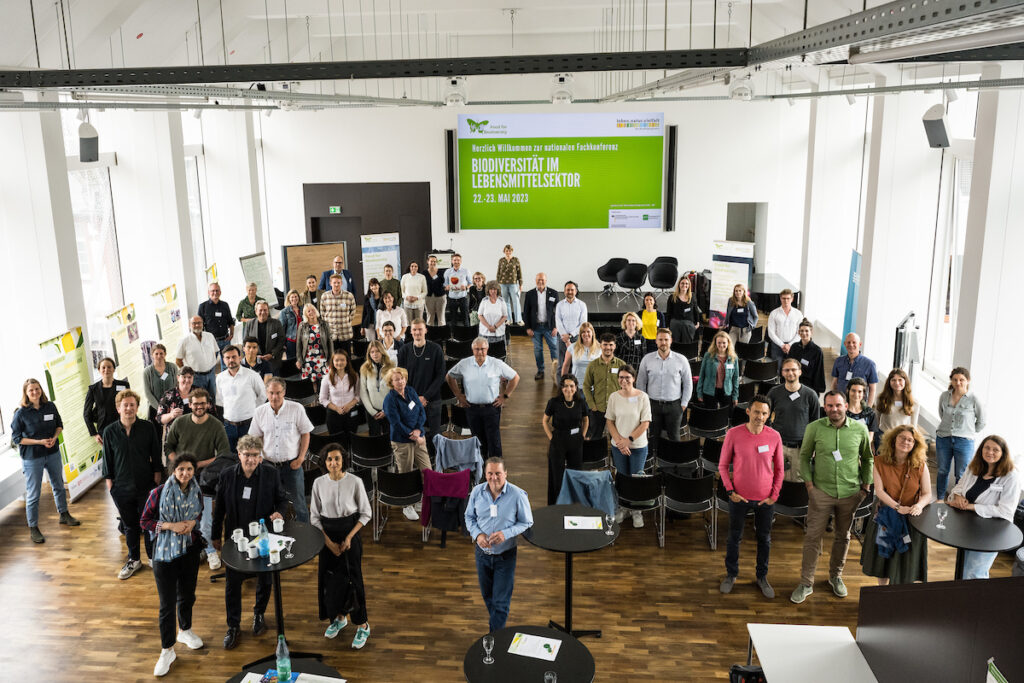 Blick in einen Konferenzsaal. Rund 50 Teilnehmer*innen stehen vor ihren Stühlen und schauen in die Kamera.