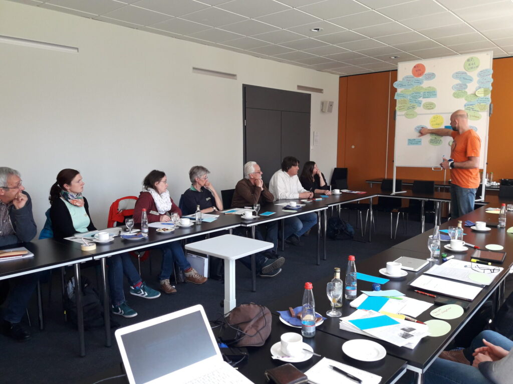 In einem Konferenzzimmer stehen schwarze Tische in U-Form. An den Tischen sitzen Menschen mit Kaffeetassen und Wasserflaschen sowie Schreibmaterial vor sich. Sie schauen auf den Mann, der vorne Moderationskärtchen an einer Pinwand befestigt.