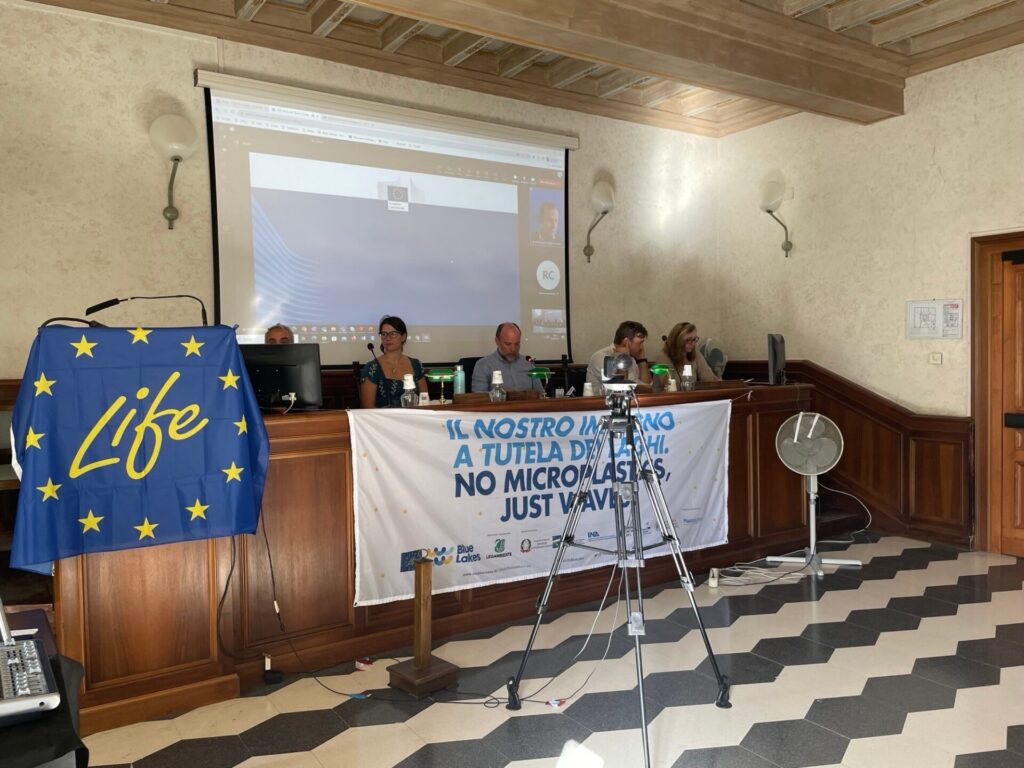 In einem Konfernzraum sitzen Menschen auf dem Podium. Vor ihnen hängt die Europa-Flagge und die Flagge des Projekts Blue Lakes.