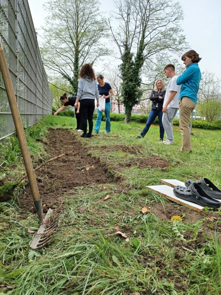 Eine Gruppe Jugendlicher steht auf einer Rasenfläche an einem Zaun. Vor ihnen ist ein Streifen der Grünfläche umgebrochen.