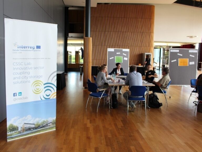 Blick in einen Veranstaltungsraum. Links steht ein Roll-up, in der Mitte sitzen Menschen zusammen an einer Tischgruppe.