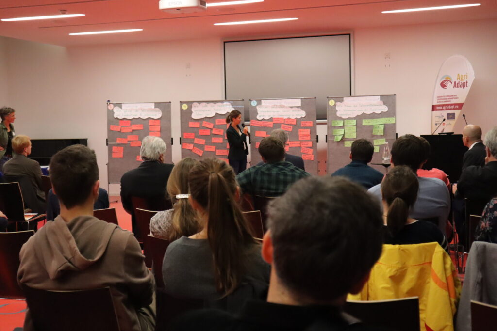 Vor einem Publikum, von dem die Hinterköpfe zu sehen sind, steht eine Referentin vor vier Pinwänden.