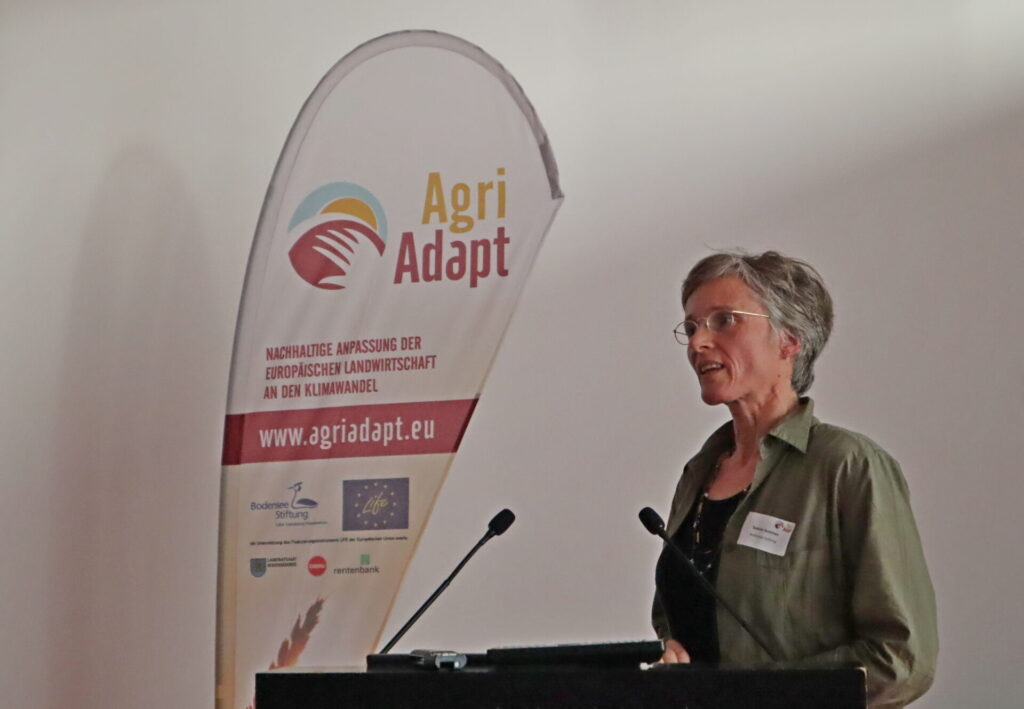 Eine Referentin steht am Rednerpult vor einer weißen Wand. Links von ihr steht eine Beachflag, auf der der Projektname AgriAdapt steht.