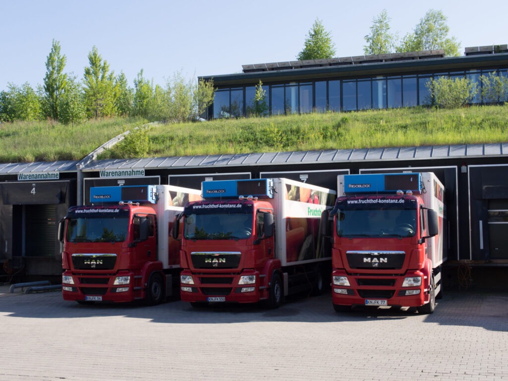 Blick auf ein Gebäude mit Gründach, auf dem auch Bäume wachsen. Im Vordergrund stehen LKWs zum Beladen an dem Gebäude.