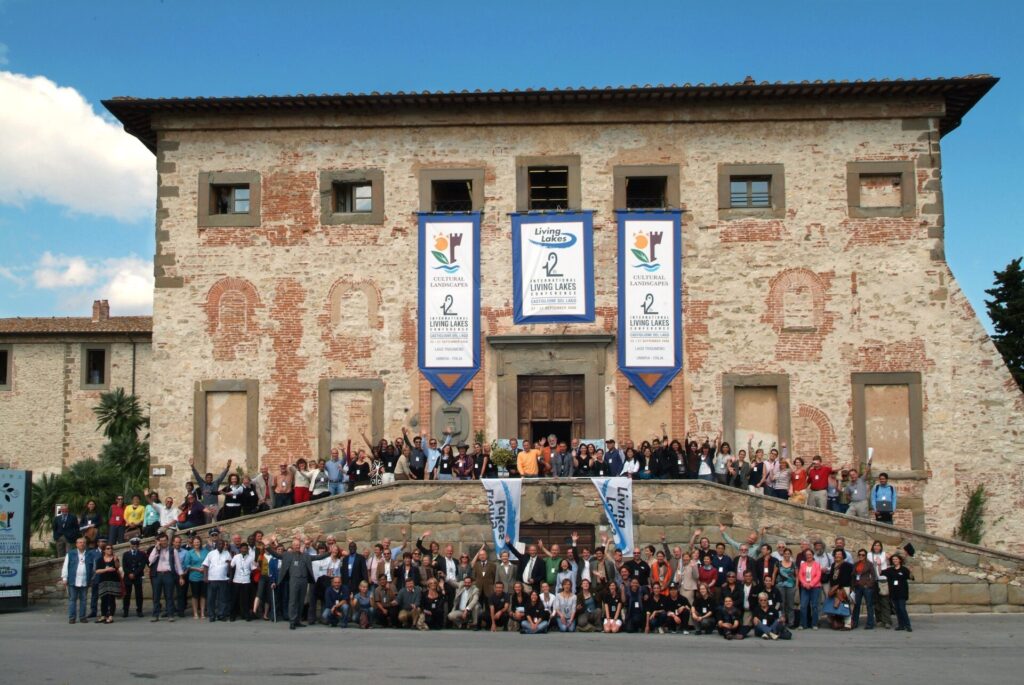 Rund 200 Menschen stehen vor einem alten Gebäude, an dessen Fassade zwei Flaggen mit der Aufschrift Living Lakes hängen.