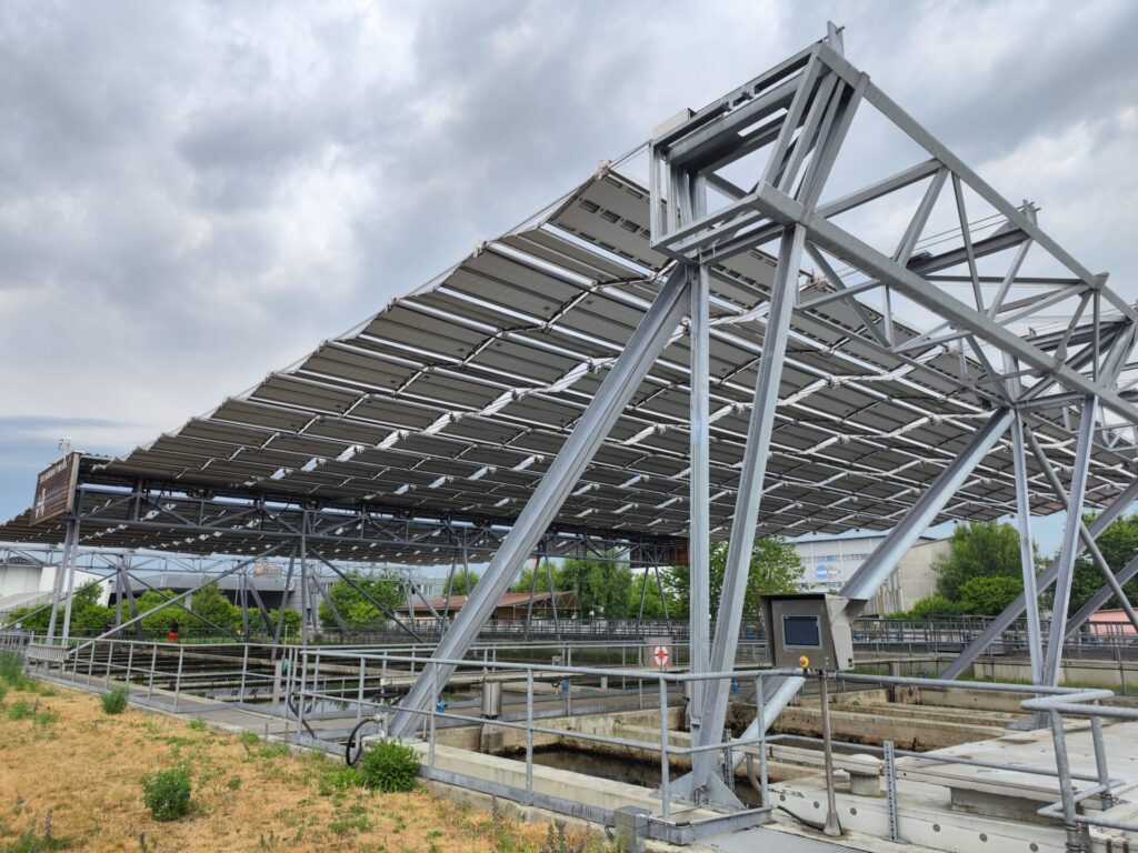 Blick von schräg unten nach oben auf die Unterseite von Solarmodulen. Unten auf dem Boden sind Becken einer Kläranlage zu erkennen,