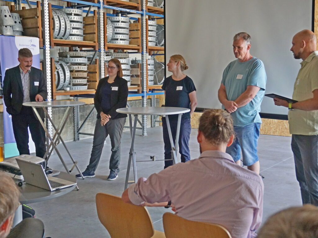 Eine Gruppe von 3 Männern und 2 Frauen steht vor Publikum an Stehtischen.