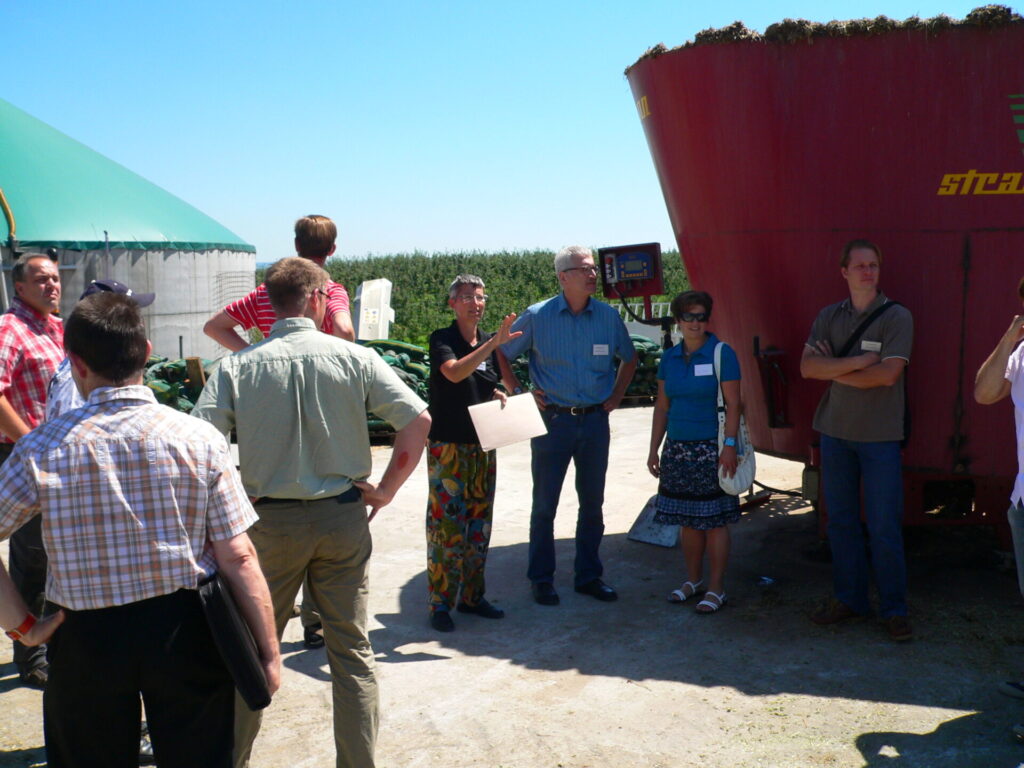 Eine Gruppe steht vor einem Maisfeld, links und rechts der Gruppe stehen Gebäude zur Verarbeitung des Mais. Eine Frau aus der Gruppe spricht.