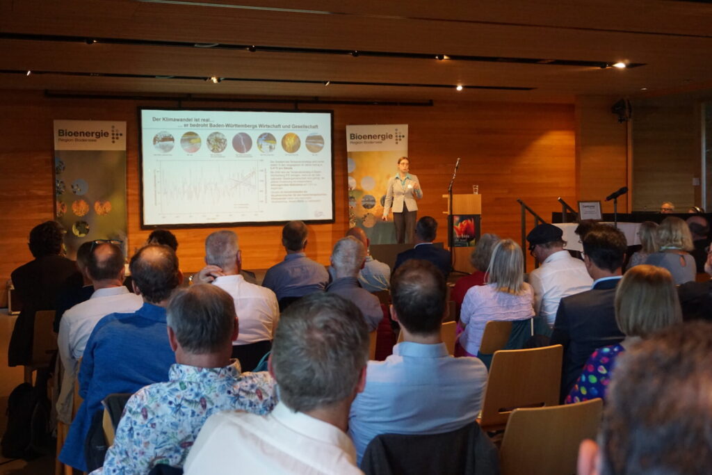 Blick von hinten in einen Vortragsraum. Das Publikum schaut auf die Referentin vorne auf der Bühne.