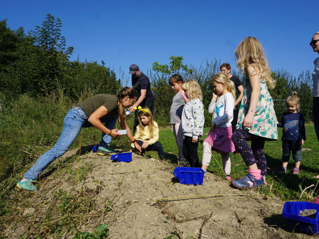 Kinder stehen an einem kleinen Hügel und schauen einer Frau zu, die Samen ausbringt.