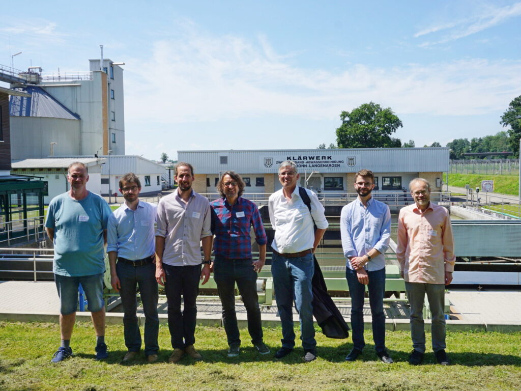 Eine Gruppe von 7 Männern steht vor einer Kläranlage und schaut in die Kamera.