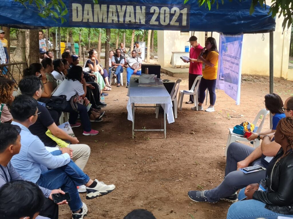 Menschen sitzen im Freien unter einem Zelt und schauen auf eine Frau mit einem Mikrofon.