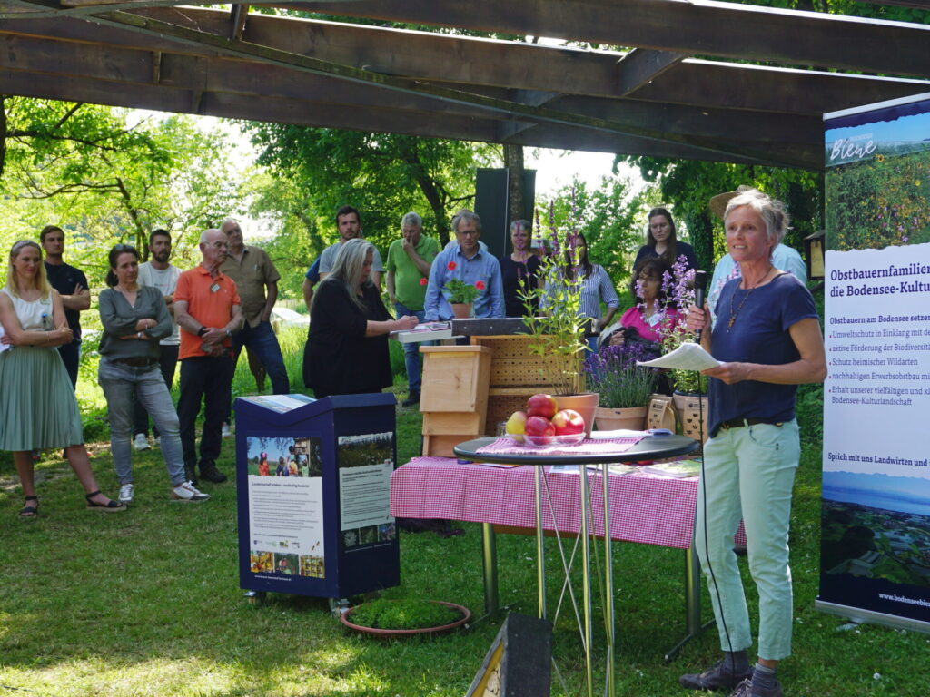 Eine Menschengruppe steht im Freien unter einem Dach und blickt auf eine Rednerin. Die Rednerin steht neben einem Tisch mit Obstprodukten.