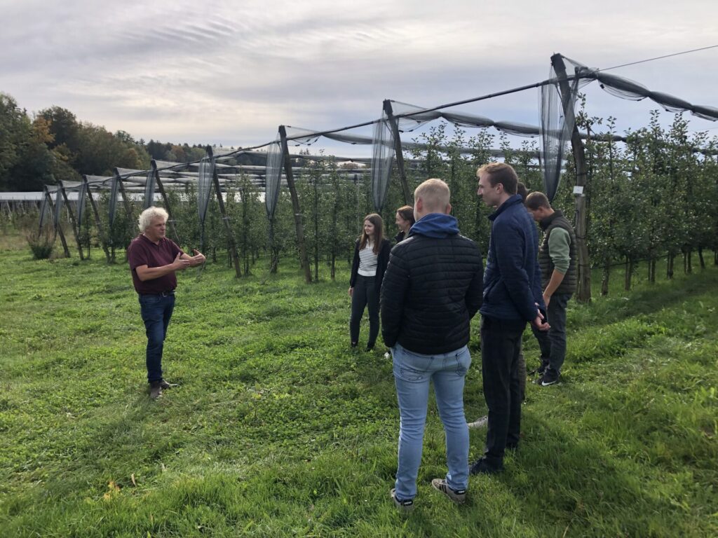 Eine Gruppe von Studierenden steht am Zeilenende einer Obstanlage. Ein Mann steht vor der Gruppe und referiert.