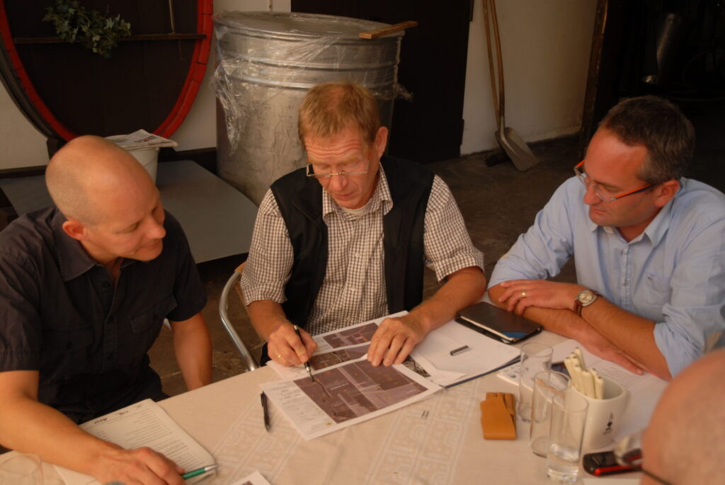 An einem Tisch sitzen drei Männer. Sie blicken auf Fotos, auf die der mittlere Mann mit einem Kugelschreiber zeigt.