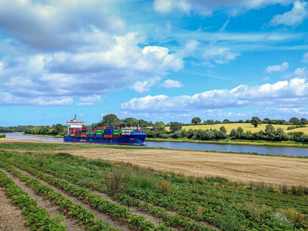 Blick auf ein Containerschiff, das inmitten von Feldern durch einen Kanal fährt.