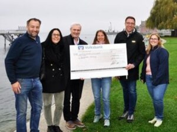 Zusehen ist Saskia Wolf mit den anderen erwähnten Personen. Sie stehen vor dem Bodensee und halten den Spenden Scheck in der Hand.