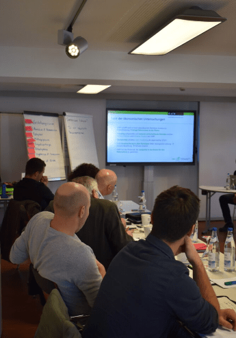 Im Blick ist ein Sitzungssaal mit großem Bildschirm vorne an der Wand. Links sind Zuhörer*innen zusehen.