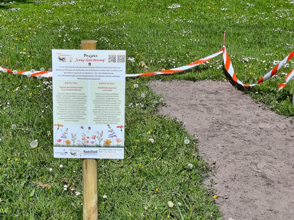 Blick auf ein Hinweisschild an einer Grünfläche. Auf der linken Fläche liegt unbearbeiteter Rasen, die rechte Fläche ist brach.