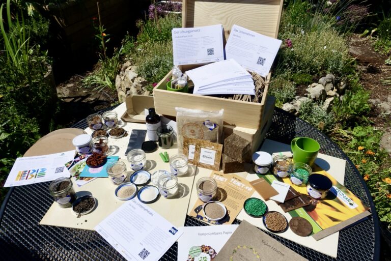 Blick auf einen Gartentisch im Grünen von oben. Auf dem Tisch steht eine geöffnete Kiste, rundherum geöffnete Gläschen mit verschiedenen bunten Inhalten.