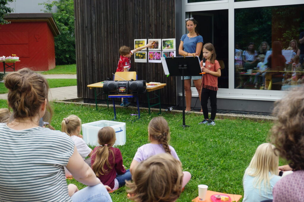 Zu sehen sind Kinder, die auf einer Wiese sitzen und nach vorne schauen. Dort steht ein kleines Mädchen mit einem Mikrofon in der Hand und liest Texte über Wildbienen vor. Links von ihr streckt ein Junge seine Hand aus und zeigt auf passende Bilder. Zwischen den beiden steht Saskia Wolf von der Bodensee-Stiftung.