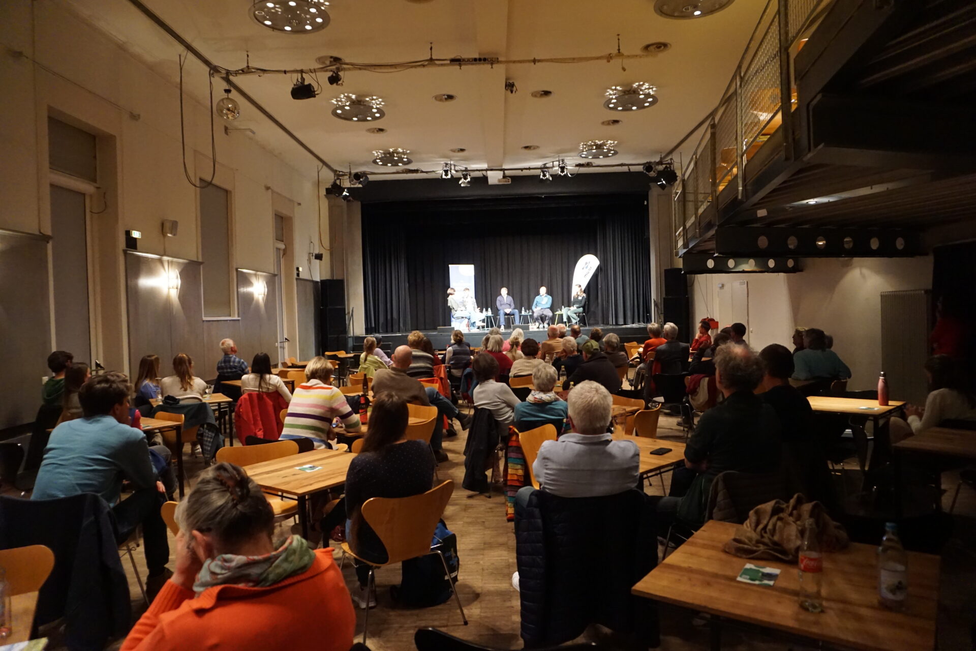 Blick von hinten in den Saal des Kulturzentrums Gems. Viele Menschen sitzen an Tischchen und blicken in Richtung Bühne, auf der fünf Menschen sitzen.