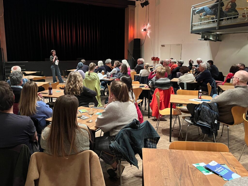 Blick von hinten in den Saal des Kulturzentrums Gems. Viele Menschen sitzen an Tischchen und blicken nach vorne auf eine Frau mit Mikrofon.