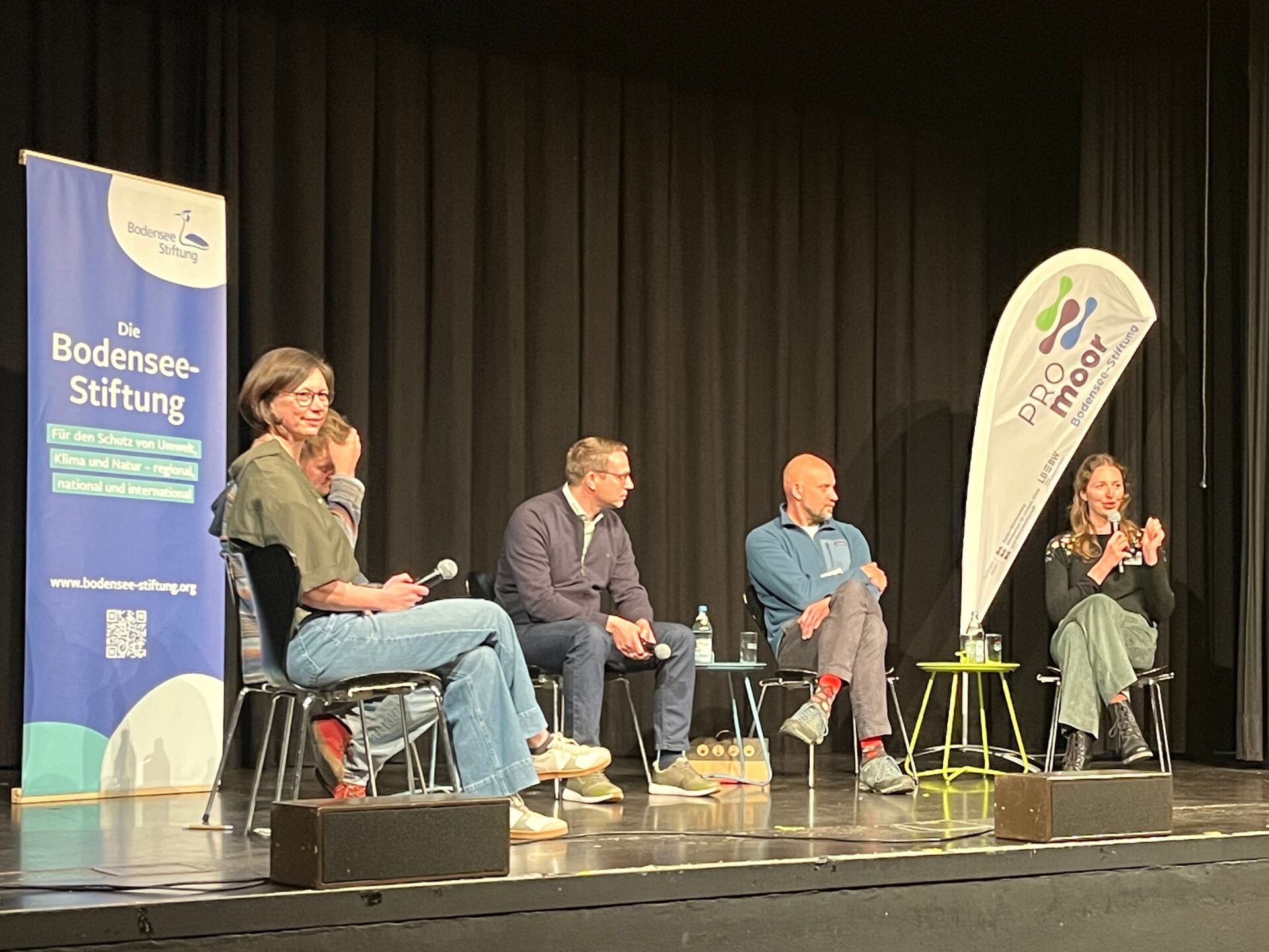 Fünf Menschen sitzen auf einer Bühne vor einem Roll-Up der Bodensee-Stiftung und einer Beachflag mit dem Logo PROmoor. Eine Frau rechts spricht in ein Mikrofon.