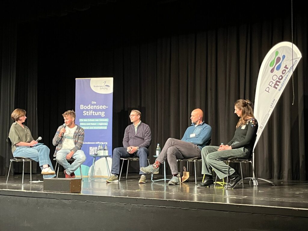 Fünf Menschen sitzen auf einer Bühne vor einem Roll-Up der Bodensee-Stiftung und einer Beachflag mit dem Logo PROmoor. Ein Mann, zweiter von links, spricht in ein Mikrofon.