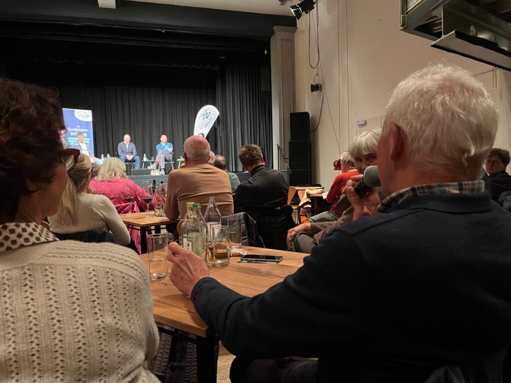 Blick durch das Publikum hindurch auf die Bühne. Rechts im Bild ist ein Mann aus dem Publikum zu sehen, der ein Mikrofon hält. 