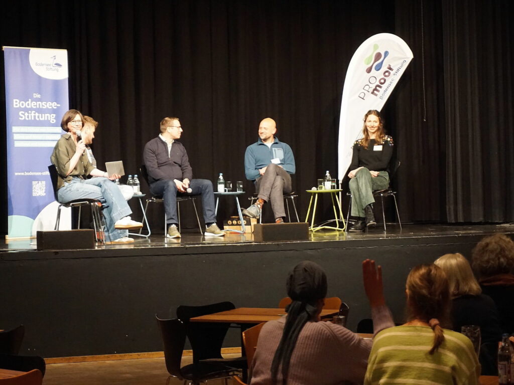 Blick auf eine Bühne, auf der fünf Menschen vor einem Roll-up und einer Beachflag sitzen. Auf dem Roll-Up steht Bodensee-Stiftung, auf der Beachflag Promoor.