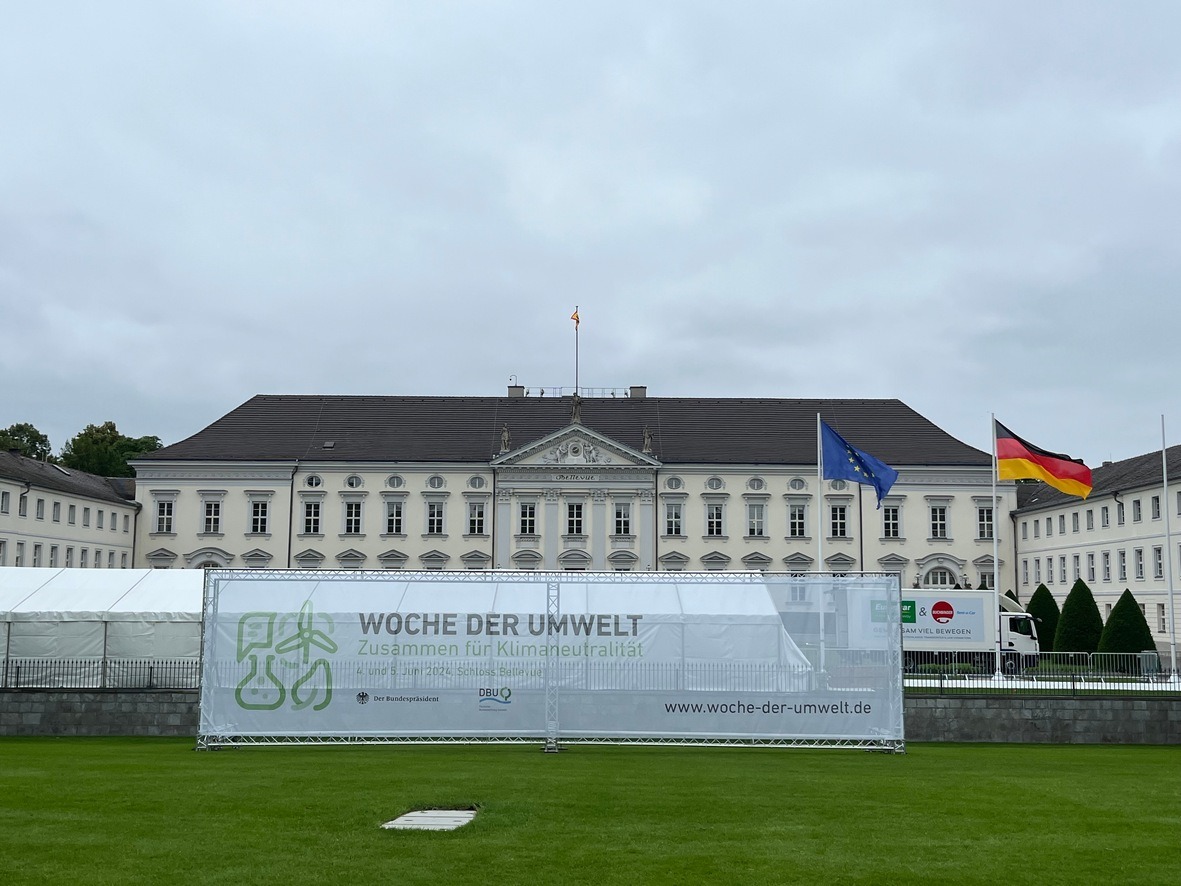 Auf dem Bild ist das Schloss Bellevue abgebildet. Rechts am Rand wehen zwei Flaggen: Die EU- und die Deutschlandflagge. Davor ist ein großer Banner mit der Aufschrift: Woche der Umwelt-Zusammen für Klimaneutralität.